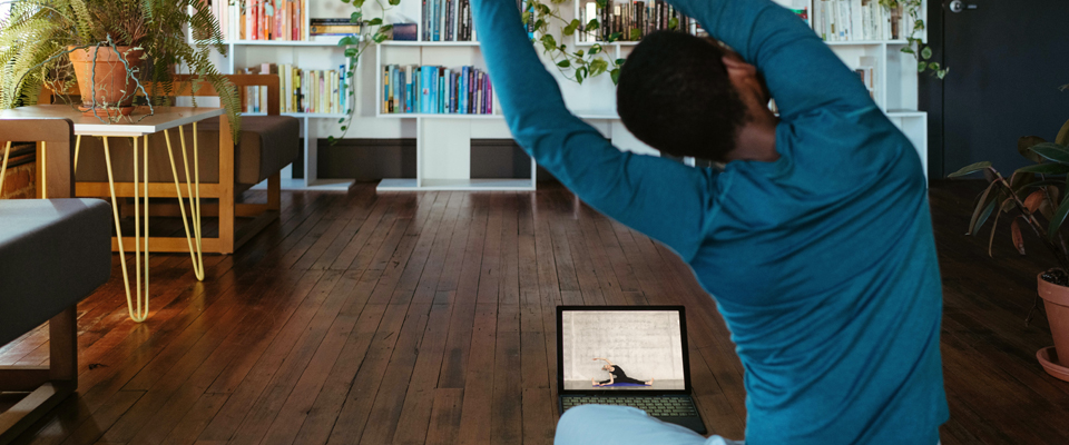 A person, seen from behind, is doing a seated yoga stretch. A laptop is on the floor in front of them, the screen shows an instructor doing the same stretch.