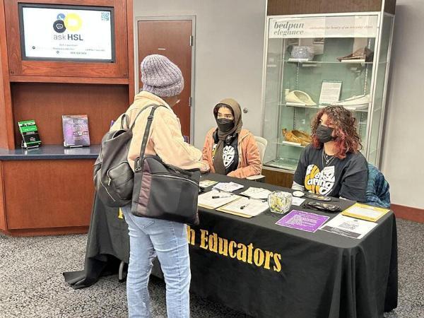 Two students sit at an information table. Another student stands in front of the table, facing them. They are having a conversation.