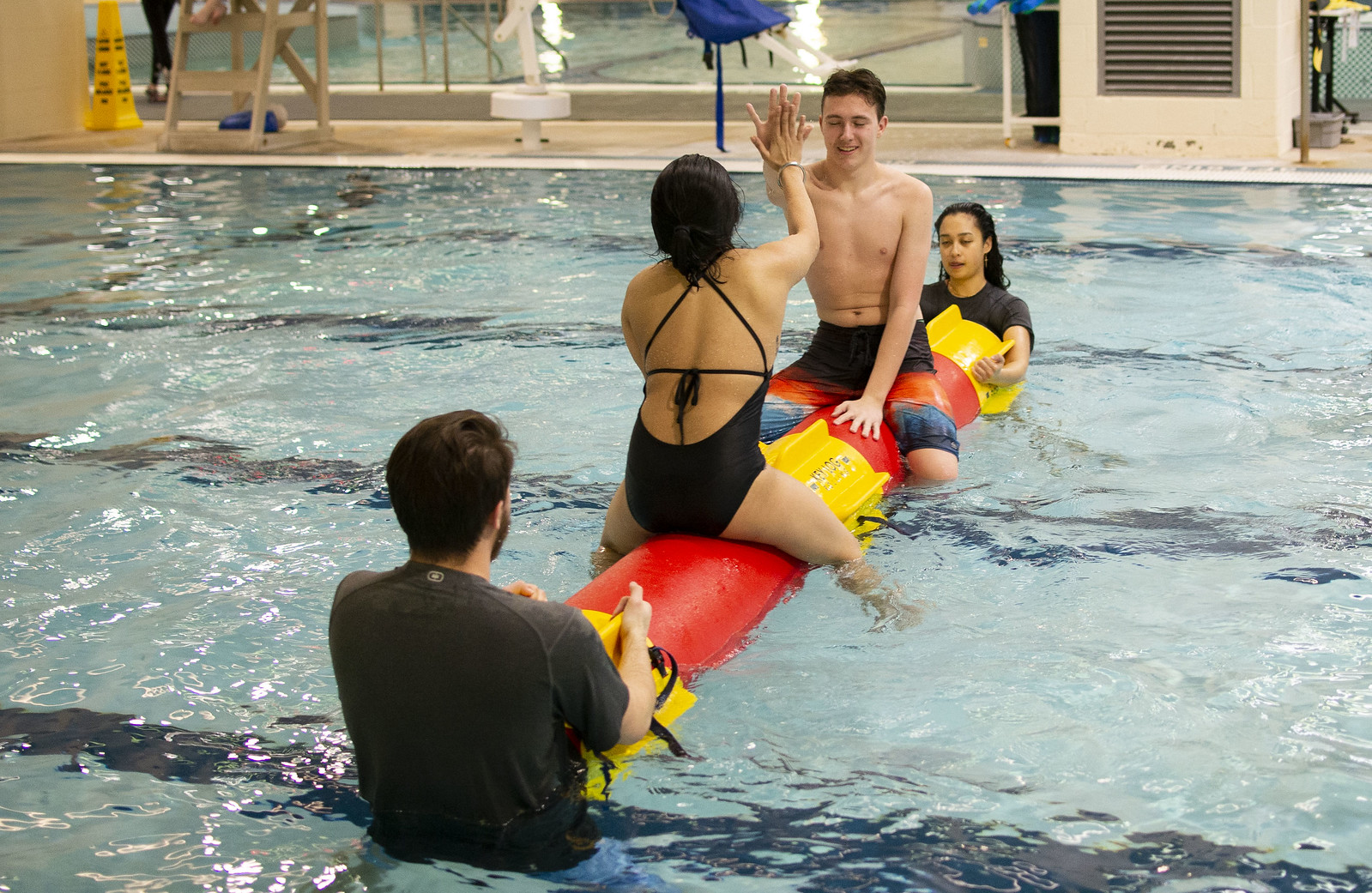 Two students high-fiving on a log in the pool while two other students hold the log steady.